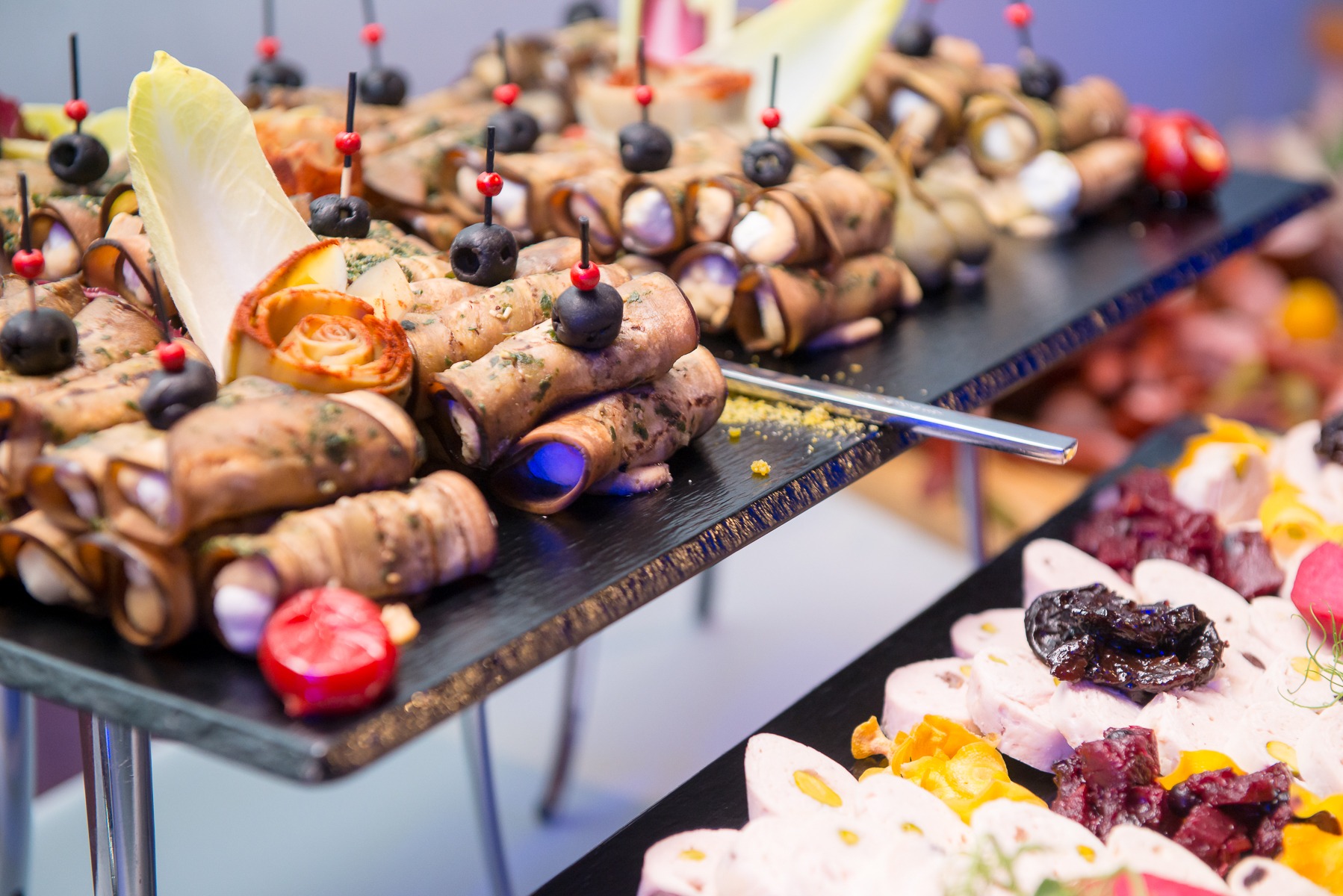 Elegancko ułożone przekąski na czarnych paterach, w tym roladki z bakłażana z oliwkami i szaszłyki z wędliną, na srebrnych stojakach, oświetlone subtelnym światłem.