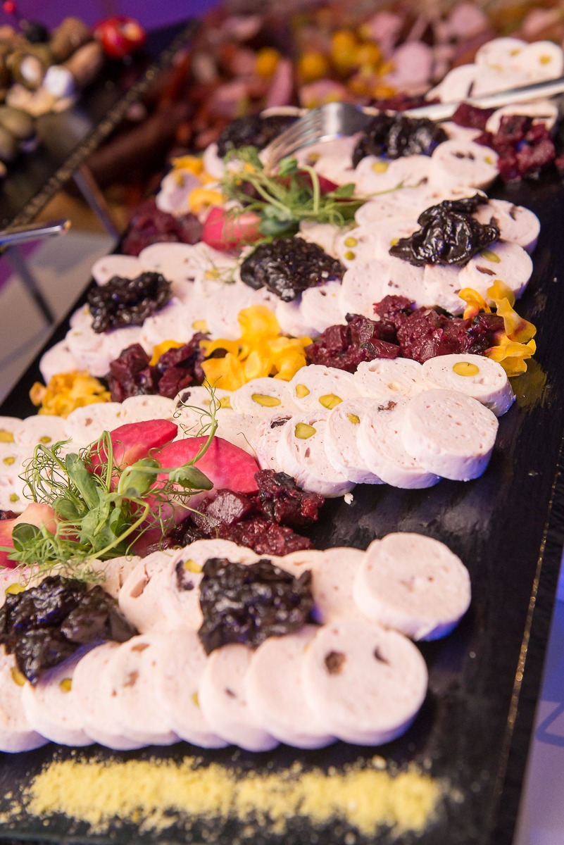 Elegancko ułożone plastry jasnej wędliny z pistacjami, udekorowane śliwkami suszonymi, buraczkami i jadalnymi kwiatami na czarnym kamiennym półmisku.