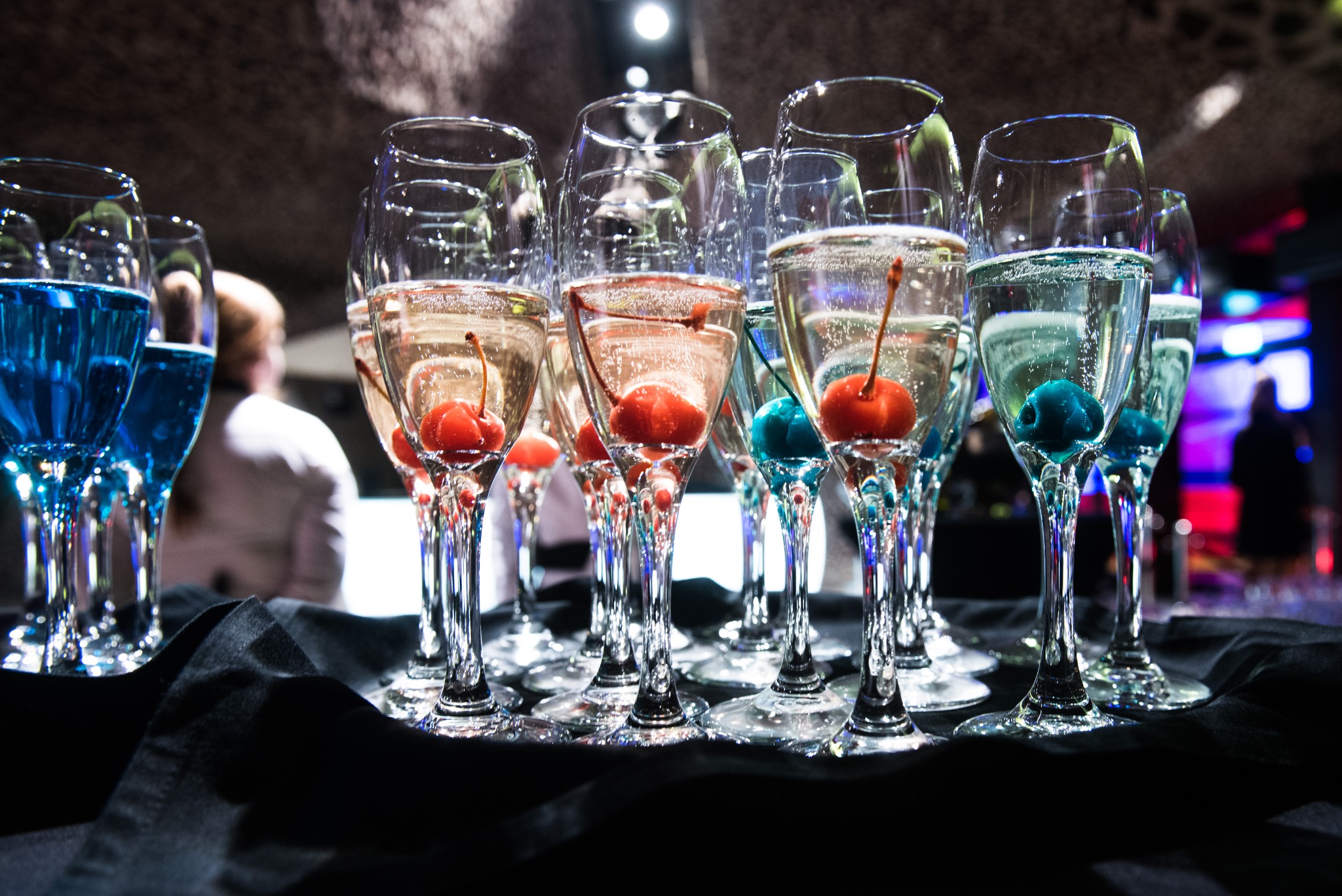 Rząd kieliszków do szampana z wiśniami, część z niebieskim barwnikiem, ustawionych na czarnym materiale w ciemnym pomieszczeniu.