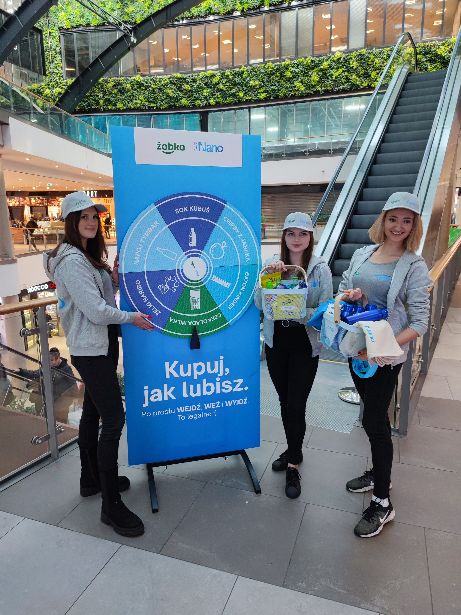 Trzy hostessy w szarych bluzach i czapkach Żabka Nano prezentują koło fortuny z nagrodami w centrum handlowym, jedna trzyma koszyk z produktami, druga koszyk z chemią, a trzecia wskazuje na koło.