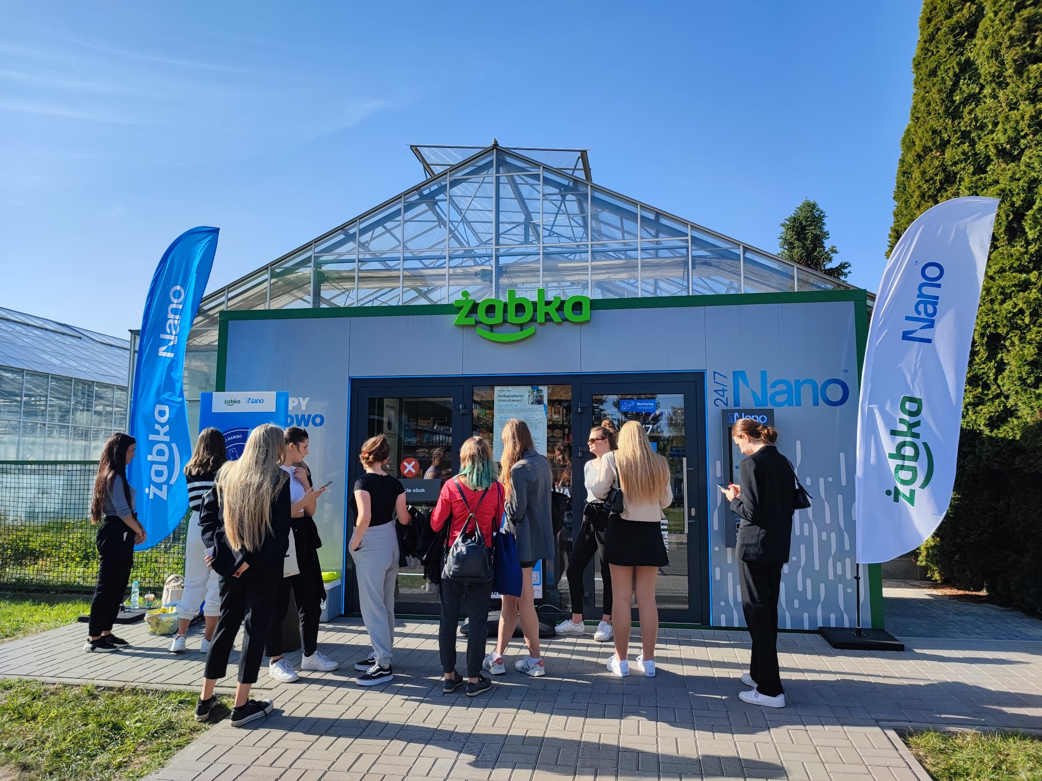 Grupa osób stoi przed sklepem Żabka Nano z przeszklonym dachem przypominającym szklarnię, widoczne flagi z logo Żabka Nano po obu stronach wejścia.