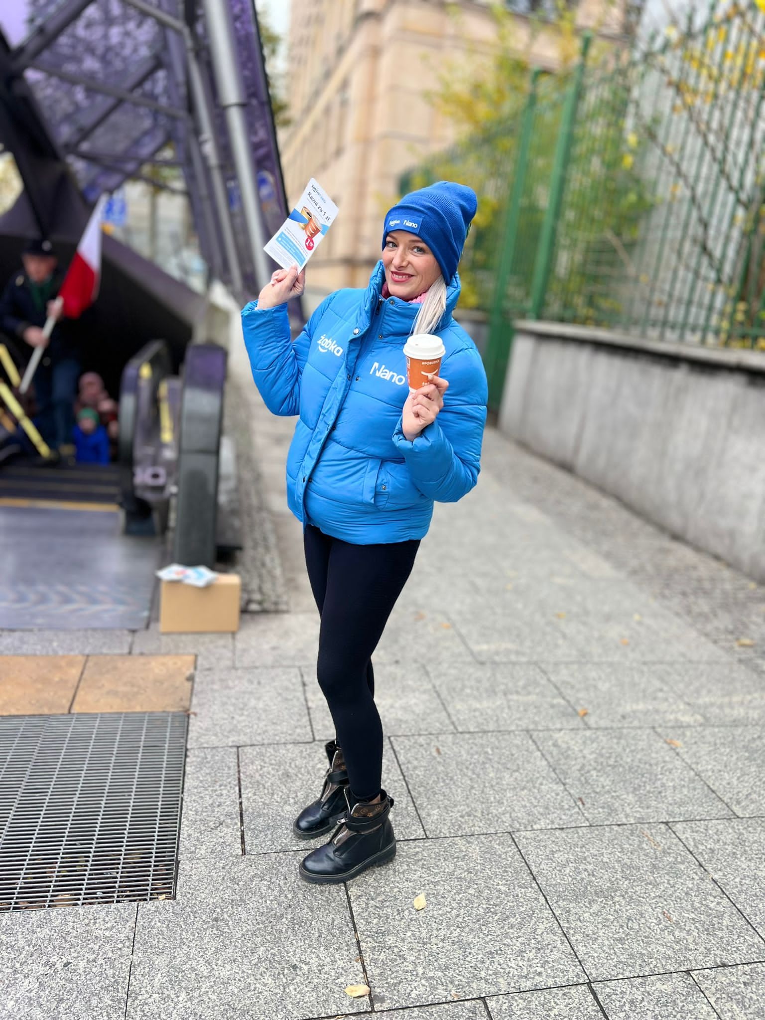 Uśmiechnięta kobieta w niebieskiej kurtce i czapce, trzymająca ulotkę i kubek z kawą przed wejściem do metra, z widocznym logo firmy na kurtce i czapce.