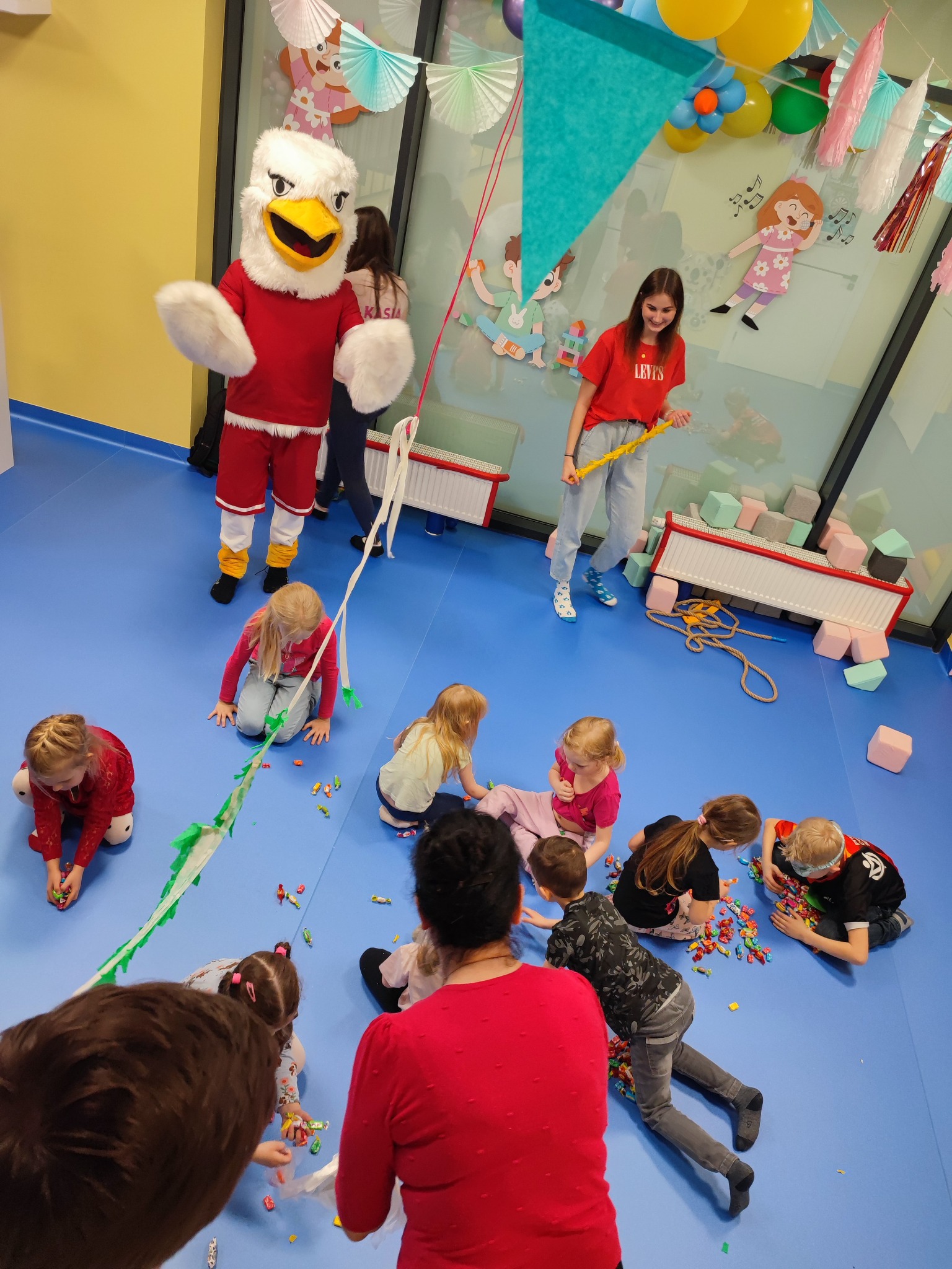 Dzieci bawiące się na podłodze podczas imprezy urodzinowej z animatorem przebranym za orła w czerwonym stroju, dekoracje wiszące na ścianach i balony.