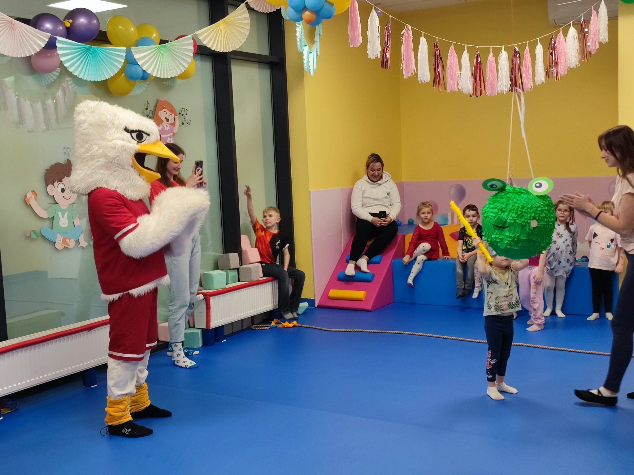 Animator w stroju orła prowadzi zabawę z piniatą dla grupy dzieci w kolorowym pokoju zabaw z dekoracjami urodzinowymi.