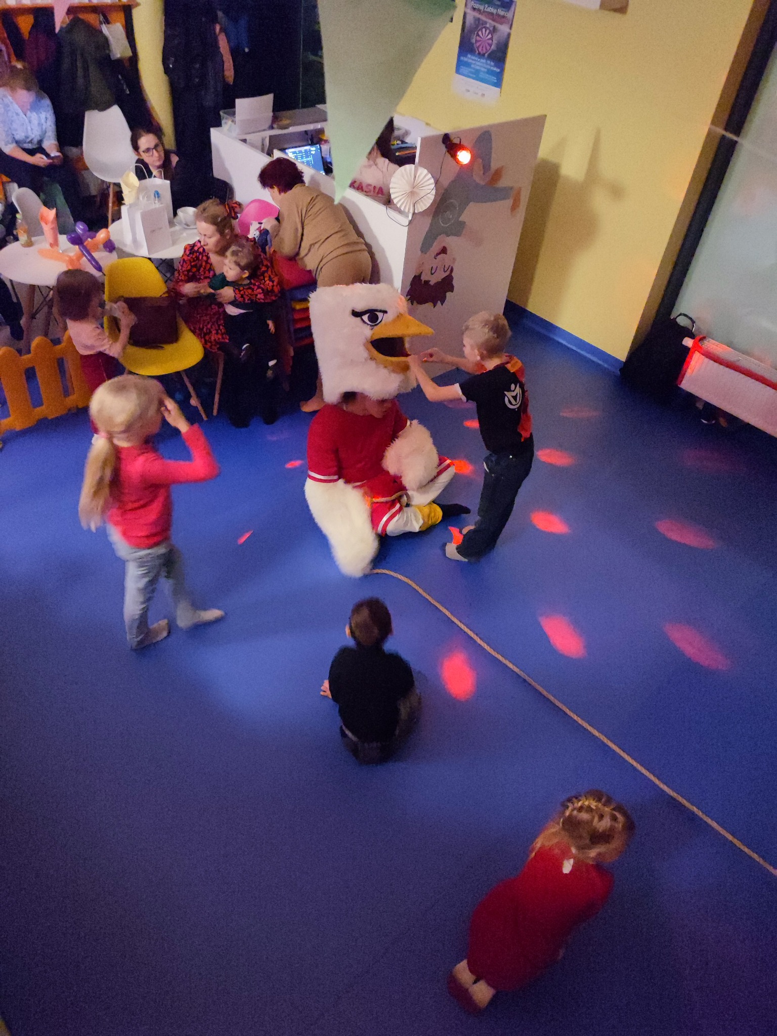 Dzieci bawią się z osobą w kostiumie orła na niebieskiej podłodze w sali zabaw. W tle siedzą rodzice przy stolikach.