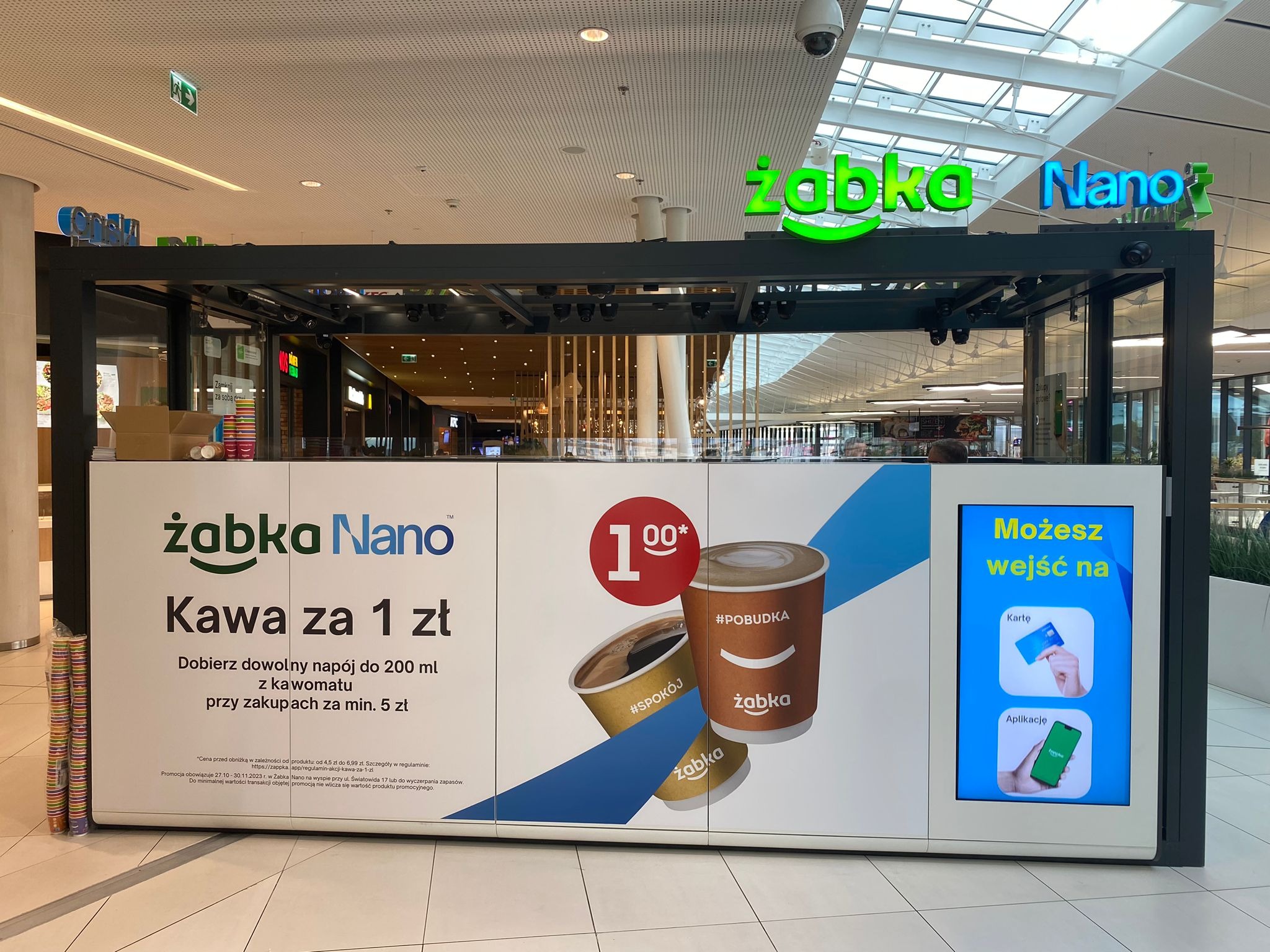 Stoisko Żabka Nano w centrum handlowym oferujące kawę za 1 zł. Widoczne logo Żabka Nano, grafika z filiżankami kawy i informacja o możliwości płatności kartą i aplikacją.