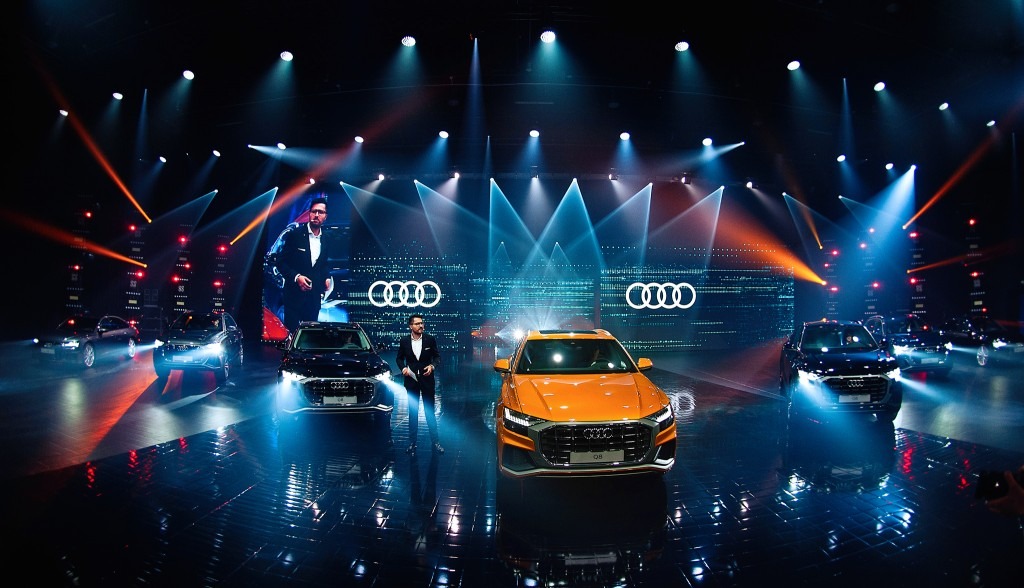 Prezentacja samochodów Audi na scenie z oświetleniem eventowym, z pomarańczowym Audi Q8 na pierwszym planie i wizerunkiem prezentera na ekranie w tle.