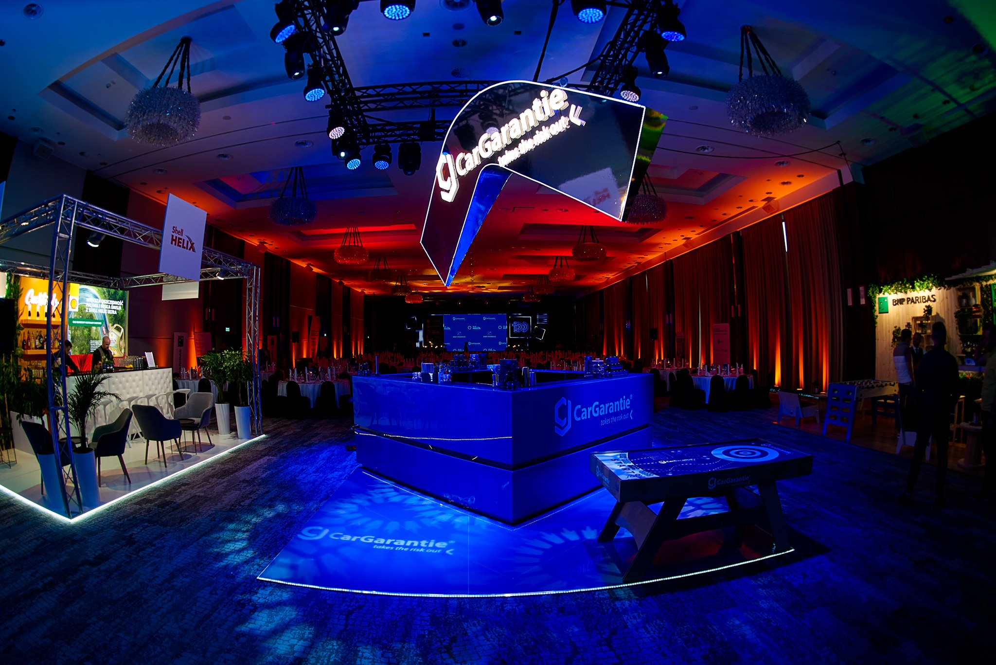 Widok ogólny na przestrzeń eventową w Warszawie z logo CarGarantie podwieszonym pod sufitem, oświetleniem punktowym i stoiskami firmowymi w tle.