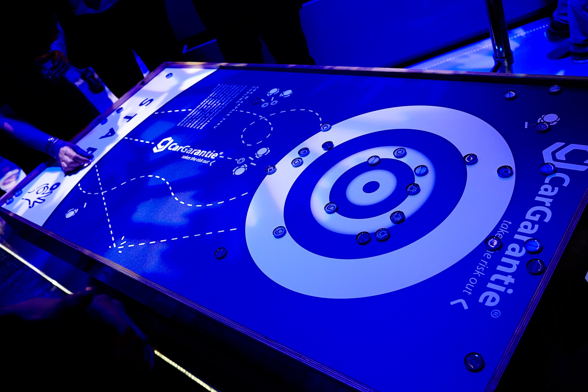 Gra curlingowa na stole z logo CarGarantie, z użyciem kapsli od butelek zamiast kamieni, oświetlona niebieskim światłem.