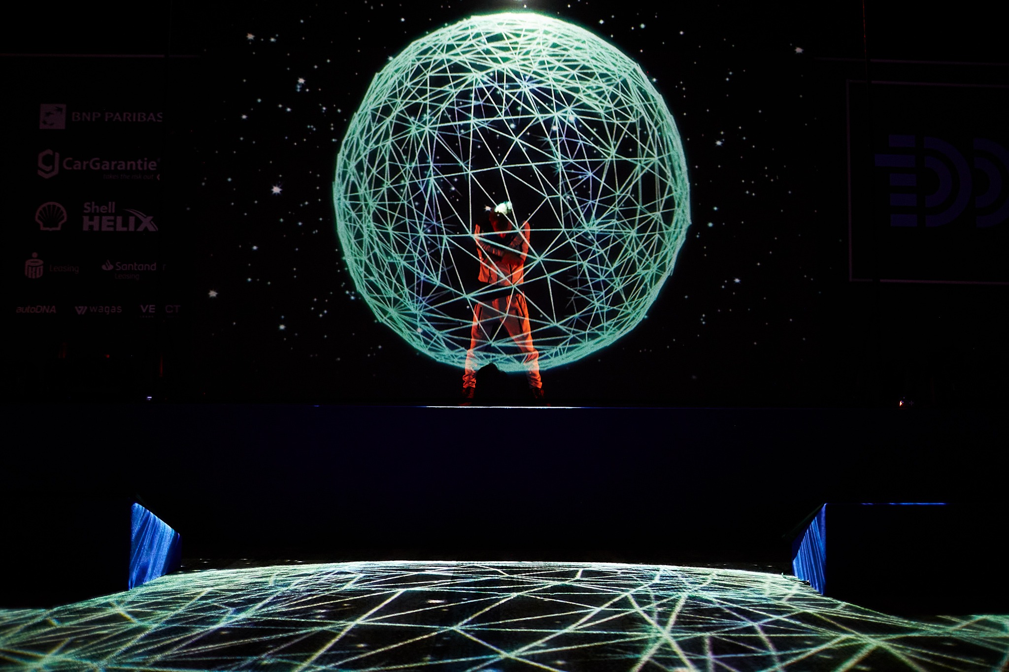 Artysta w pomarańczowym kostiumie wewnątrz trójwymiarowej, świecącej kuli na scenie z projekcją geometrycznych wzorów, logo sponsorów w tle