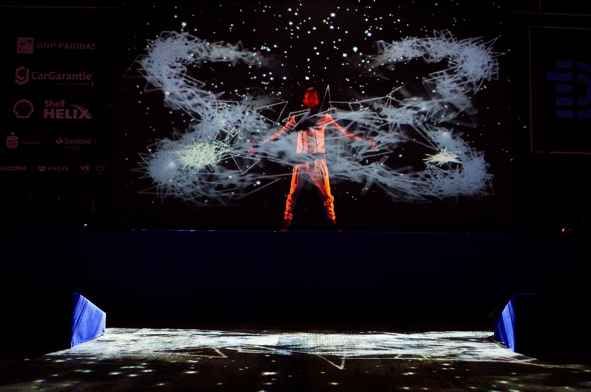 Artysta w pomarańczowym kostiumie na scenie z projekcją grafiki przypominającej konstelację, oświetlony od dołu, widoczne logotypy sponsorów w tle.