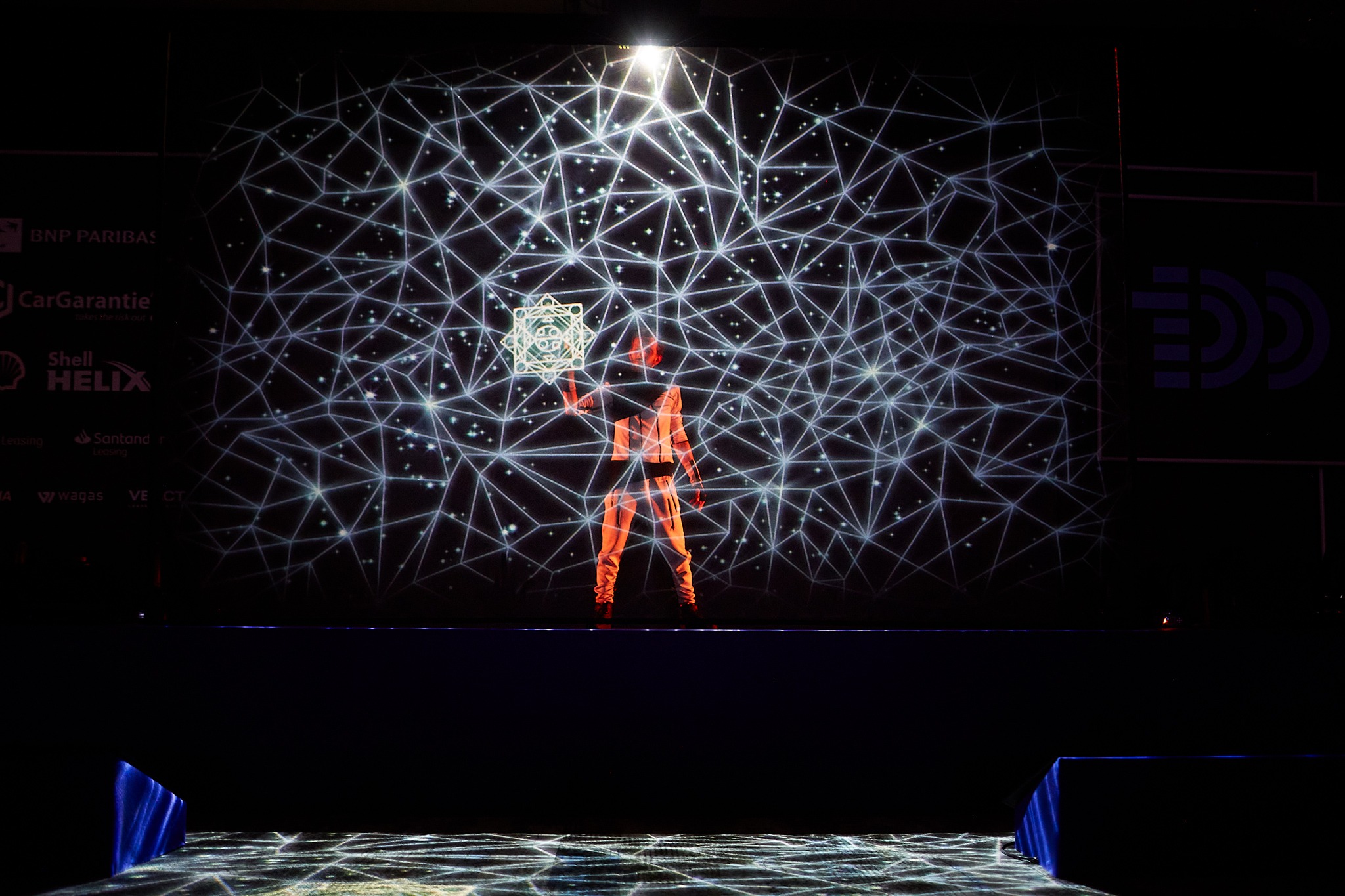 Interaktywny pokaz sceniczny: performer w pomarańczowym kostiumie z podświetlonym rekwizytem na tle geometrycznej projekcji świetlnej.