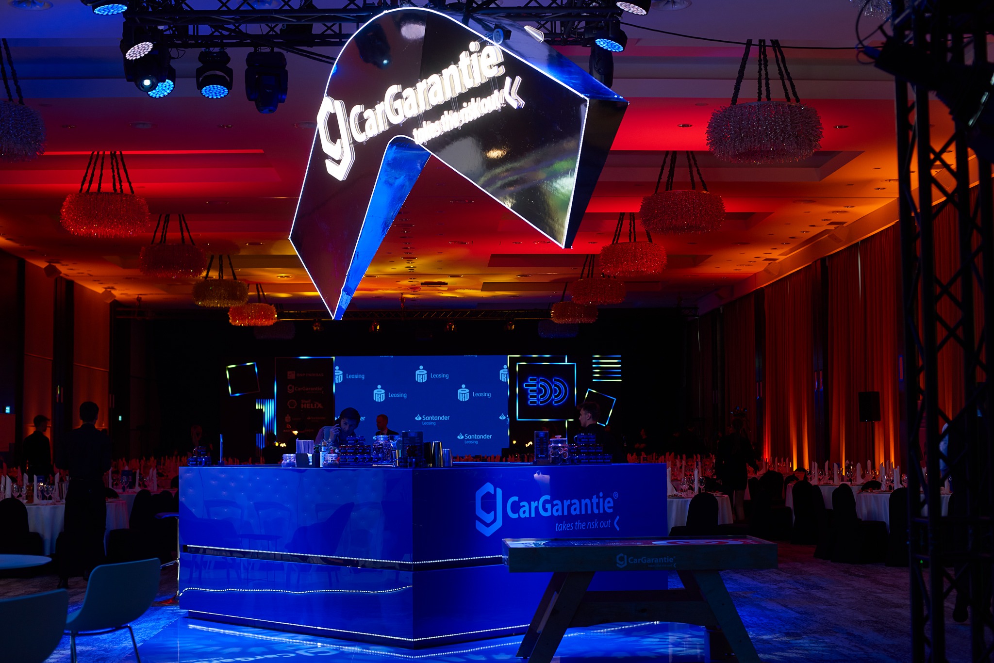 Sala konferencyjna z podświetlonym barem w kolorze niebieskim z logo firmy CarGarantie, dekoracyjne lampy wiszące pod sufitem, oświetlenie sceniczne w odcieniach czerwieni i niebieskiego.