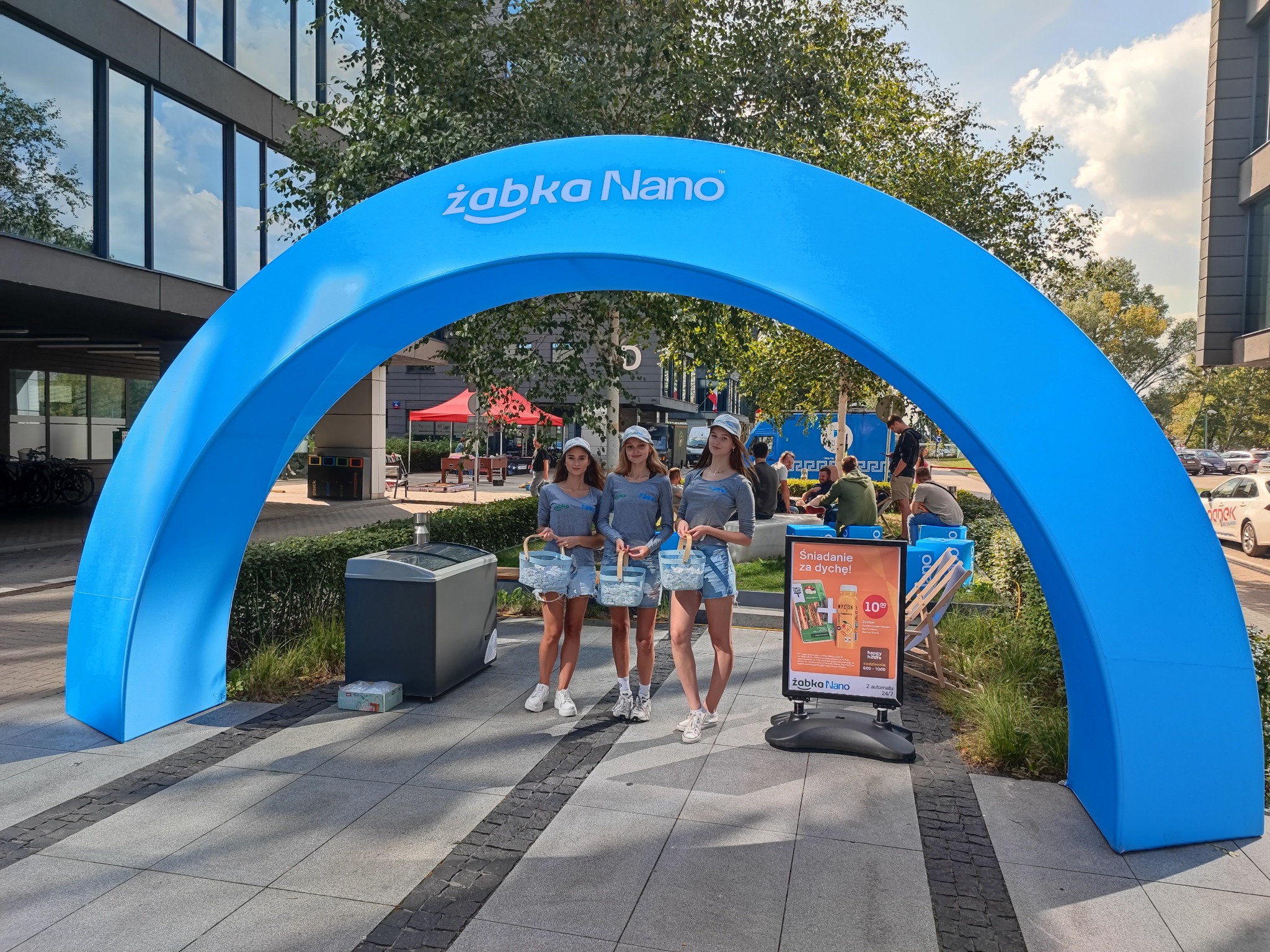 Promocja Żabka Nano z hostessami rozdającymi produkty pod niebieskim łukiem z logo, na tle budynku biurowego i drzew.