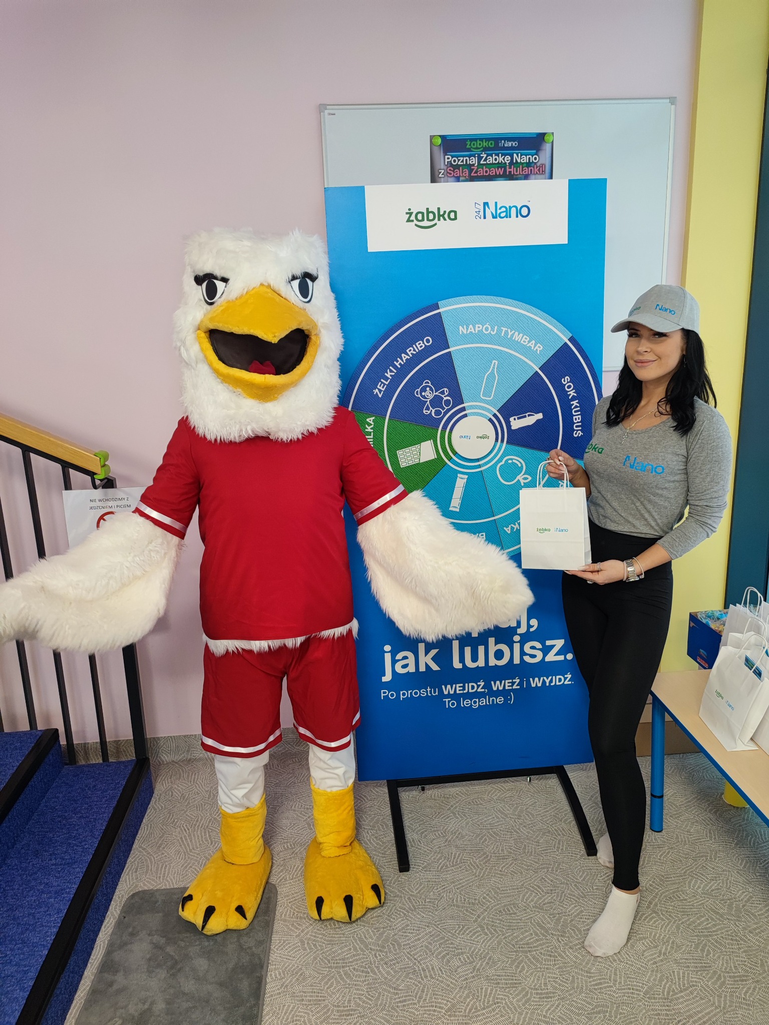 Mascot orła w czerwonym stroju sportowym pozuje obok kobiety w szarej koszulce i czarnych legginsach, trzymającej białą torbę z logo Żabka Nano, na tle plakatu reklamowego sieci sklepów Żabka Nano.