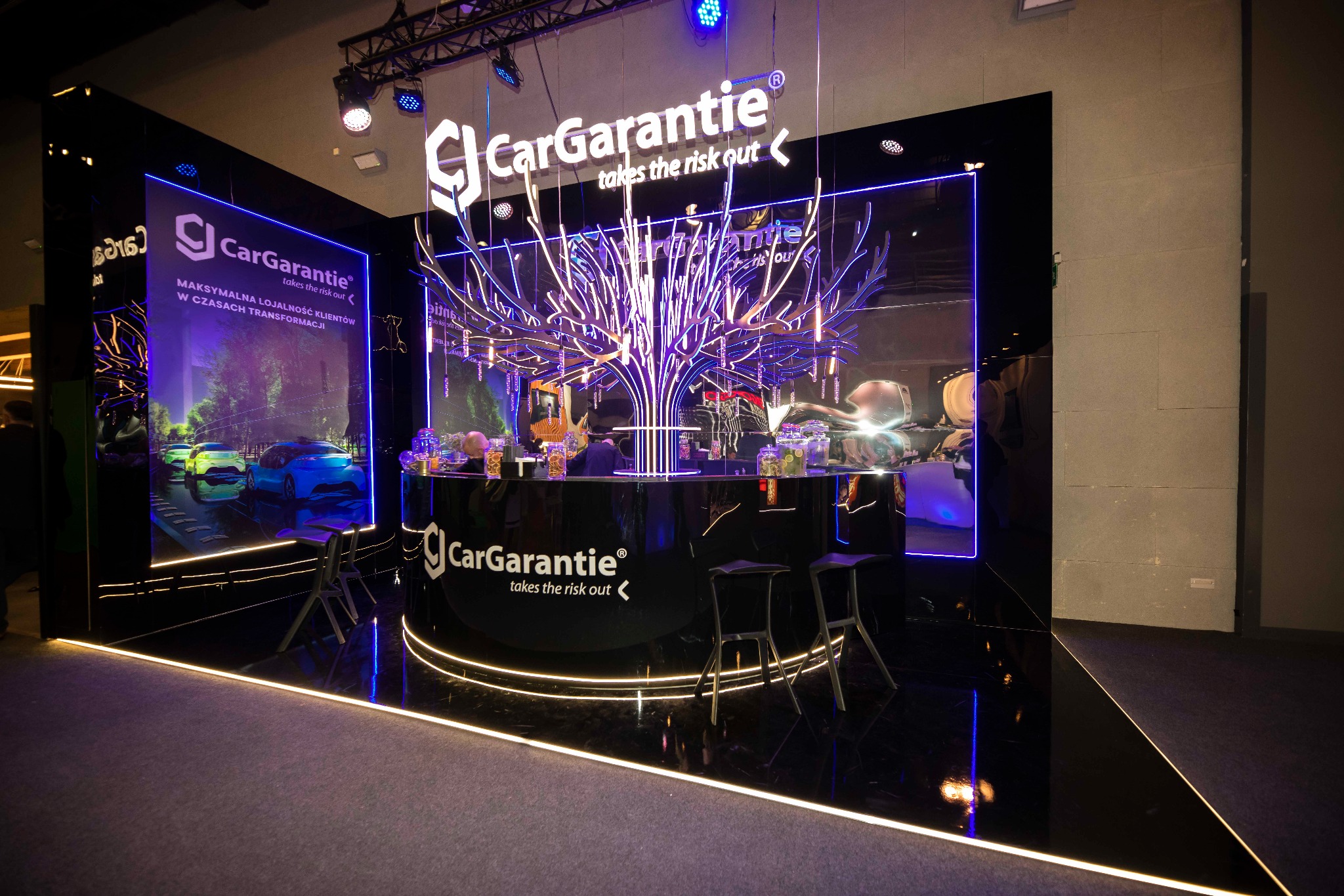 Stoisko targowe CarGarantie z podświetlanym drzewem z neonów, czarnym barem i krzesłami, oraz ekranem z samochodami w tle.
