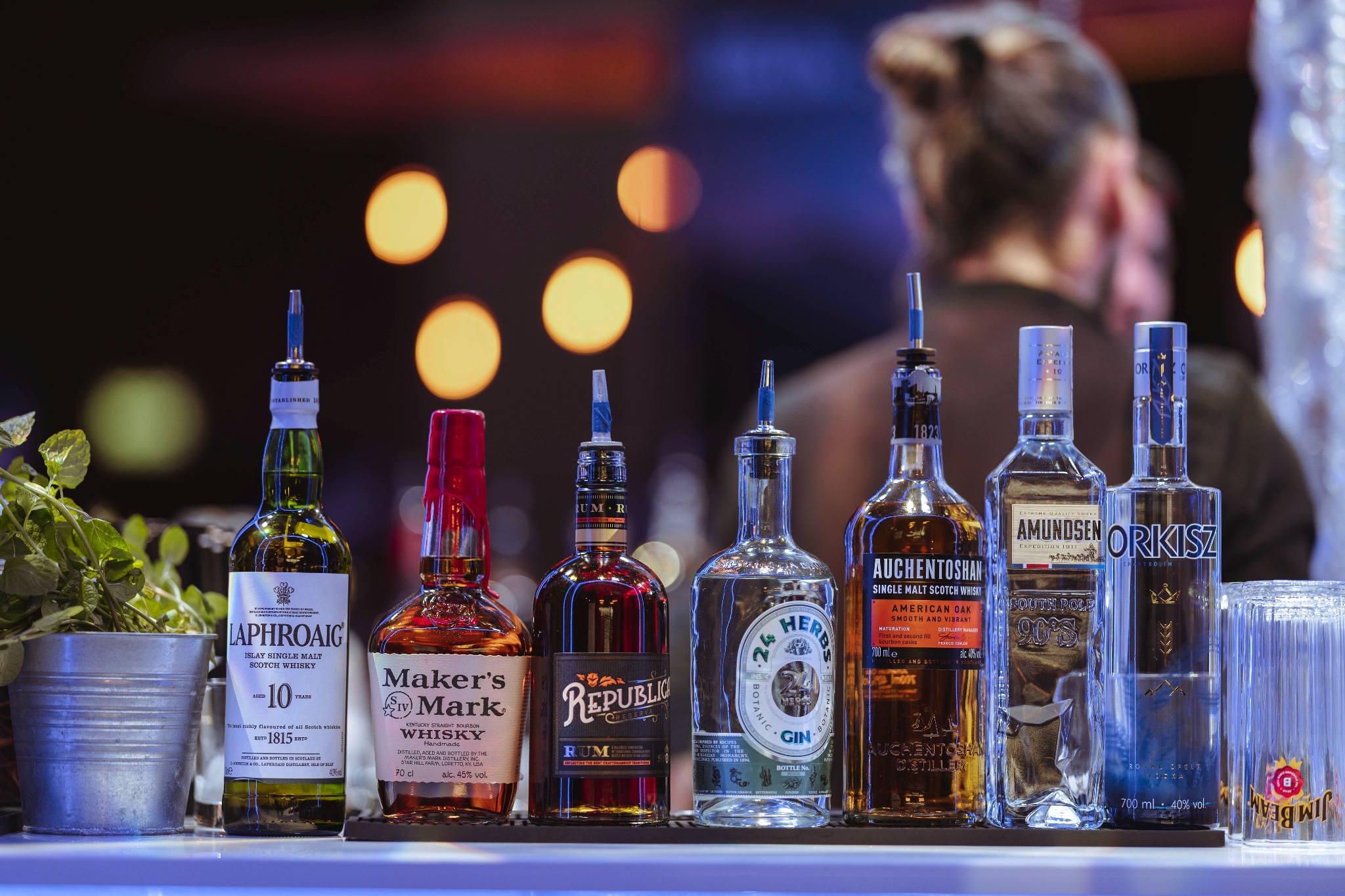 Rząd butelek różnych alkoholi, w tym whisky Laphroaig i Maker's Mark, rum Republica, gin 24 Herbs, wódka Amundsen i Orkisz, na barze z dekoracyjnym oświetleniem w tle i zielenią w metalowym...