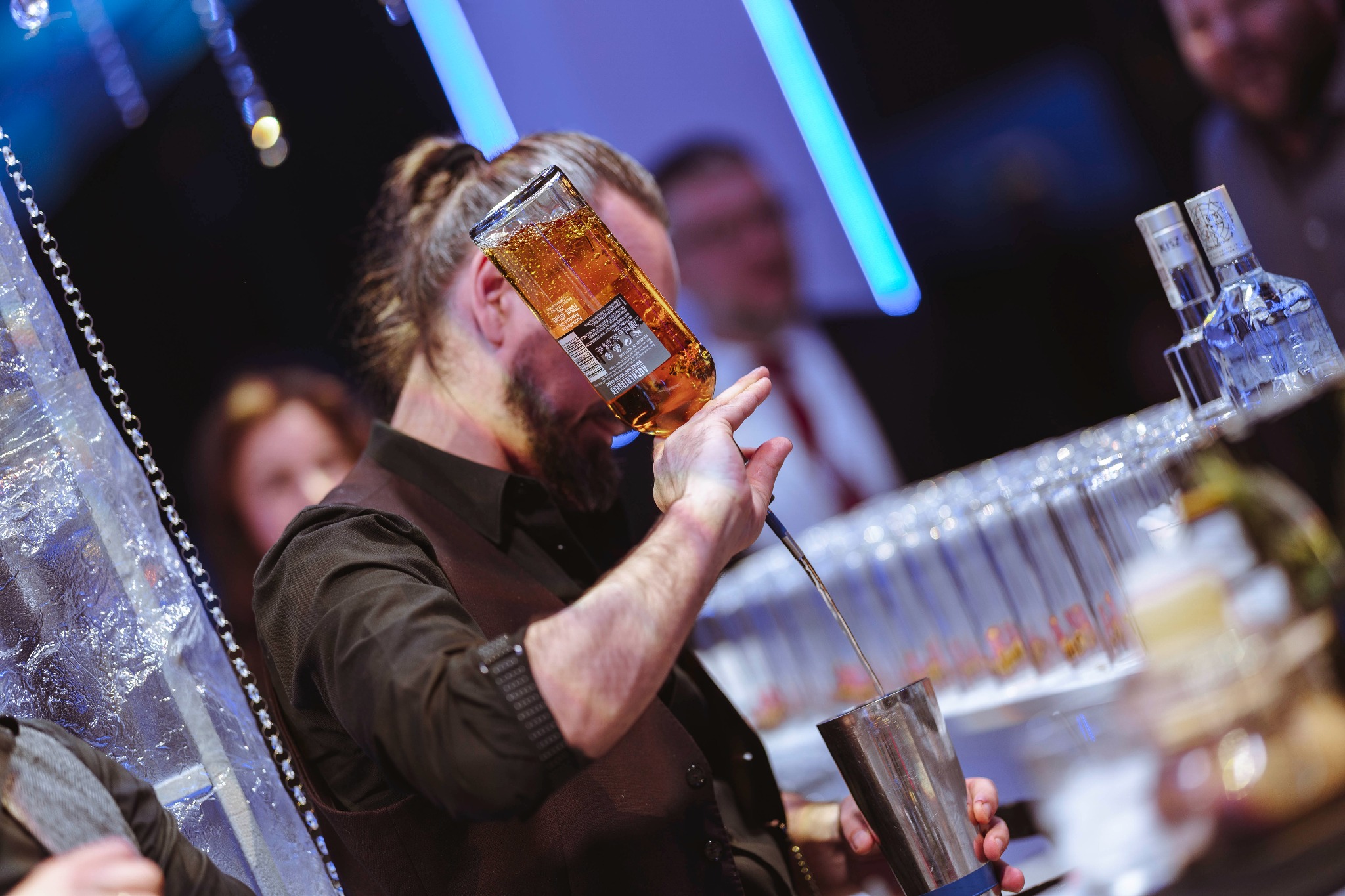 Barmen w brązowej kamizelce nalewa bursztynowy płyn z butelki Auchentoshan do shakera na tle rzędu kieliszków i dekoracji z lodu podczas eventu.
