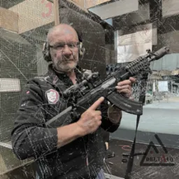 Mężczyzna w okularach i słuchawkach ochronnych prezentuje karabin AK-47 z lunetą na strzelnicy, widoczne logo Delta 10 w rogu.