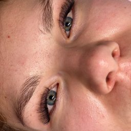 BellaLashes.pl - Stylizacja & Przedłużanie Rzęs Pniewy - Zbliżenie na twarz kobiety po zabiegu przedłużania rzęs, z wyraźnie widocznymi, gęstymi, sztucznymi rzęsami i naturalnymi brwiami, na różowym podkładzie.