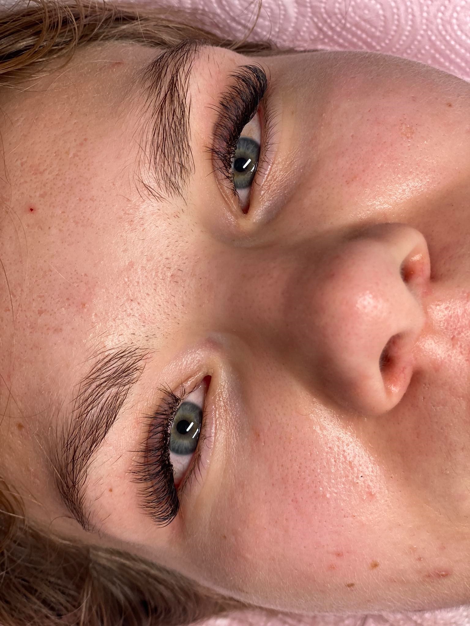 Zbliżenie na twarz kobiety po zabiegu przedłużania rzęs, z wyraźnie widocznymi, gęstymi, sztucznymi rzęsami i naturalnymi brwiami, na różowym podkładzie.