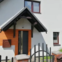Dom jednorodzinny z białą elewacją i fragmentem imitującym drewno, ciemna stolarka okienna i zadaszenie nad wejściem, skrzynka gazowa w kolorze elewacji.