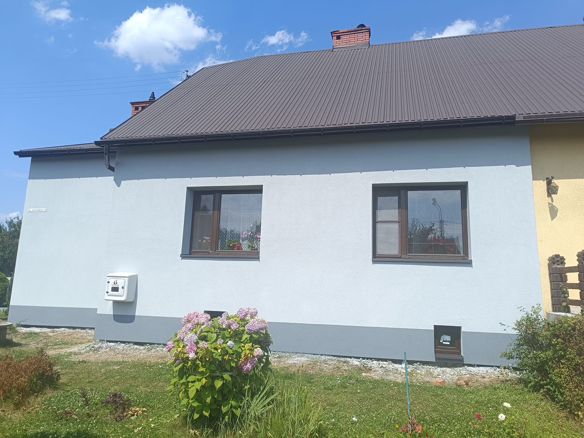 Dom jednorodzinny z szarą elewacją, brązowym dachem i oknami, widoczny fragment żółtej ściany sąsiedniego budynku, na pierwszym planie krzew hortensji.