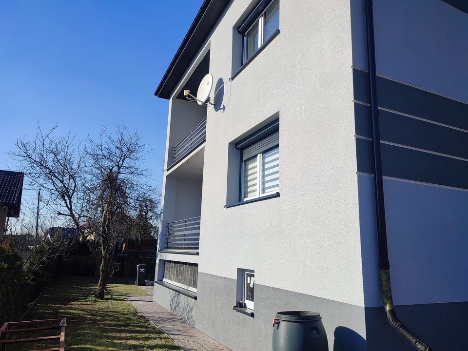 Dwukondygnacyjny budynek z szarą elewacją, balkonem i anteną satelitarną, widziany z poziomu ogrodu w słoneczny dzień.