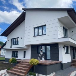 Ceglak - Nowoczesny dom jednorodzinny z białą elewacją, ciemnymi oknami i wejściem z brązowymi schodami, na których stoi aluminiowa drabina, numer domu 145.