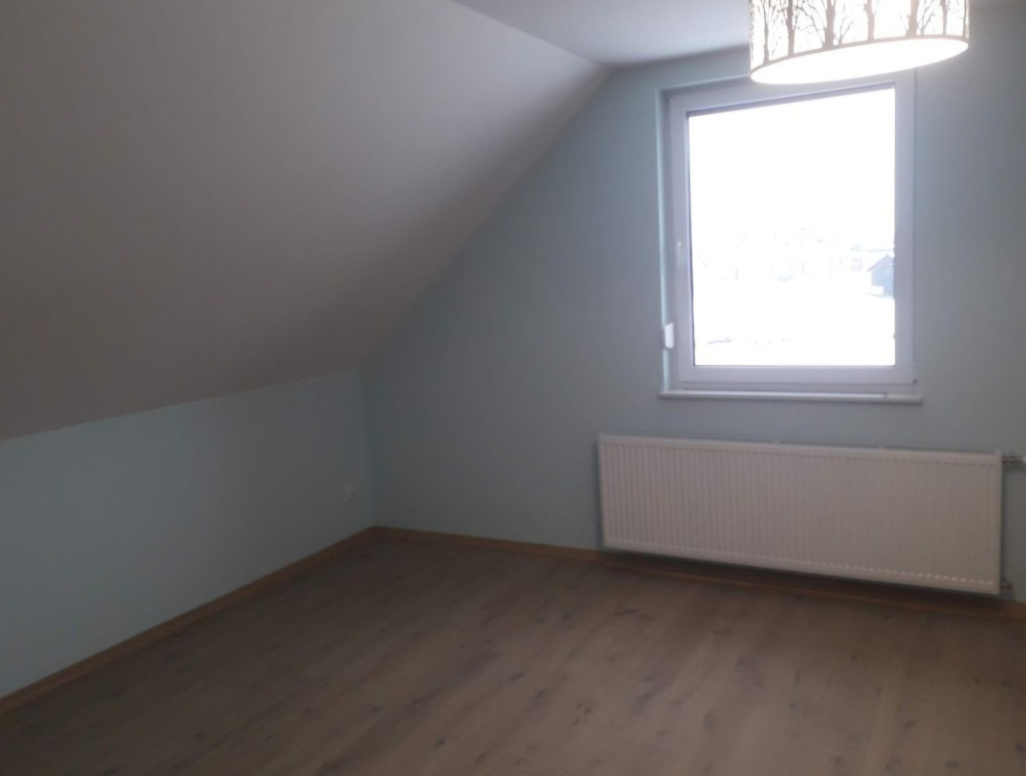 Puste poddasze po remoncie: świeżo pomalowane ściany w odcieniach szarości i błękitu, drewnopodobna podłoga, biały grzejnik pod oknem z widokiem na zabudowania, lampa sufitowa z motywem drzew