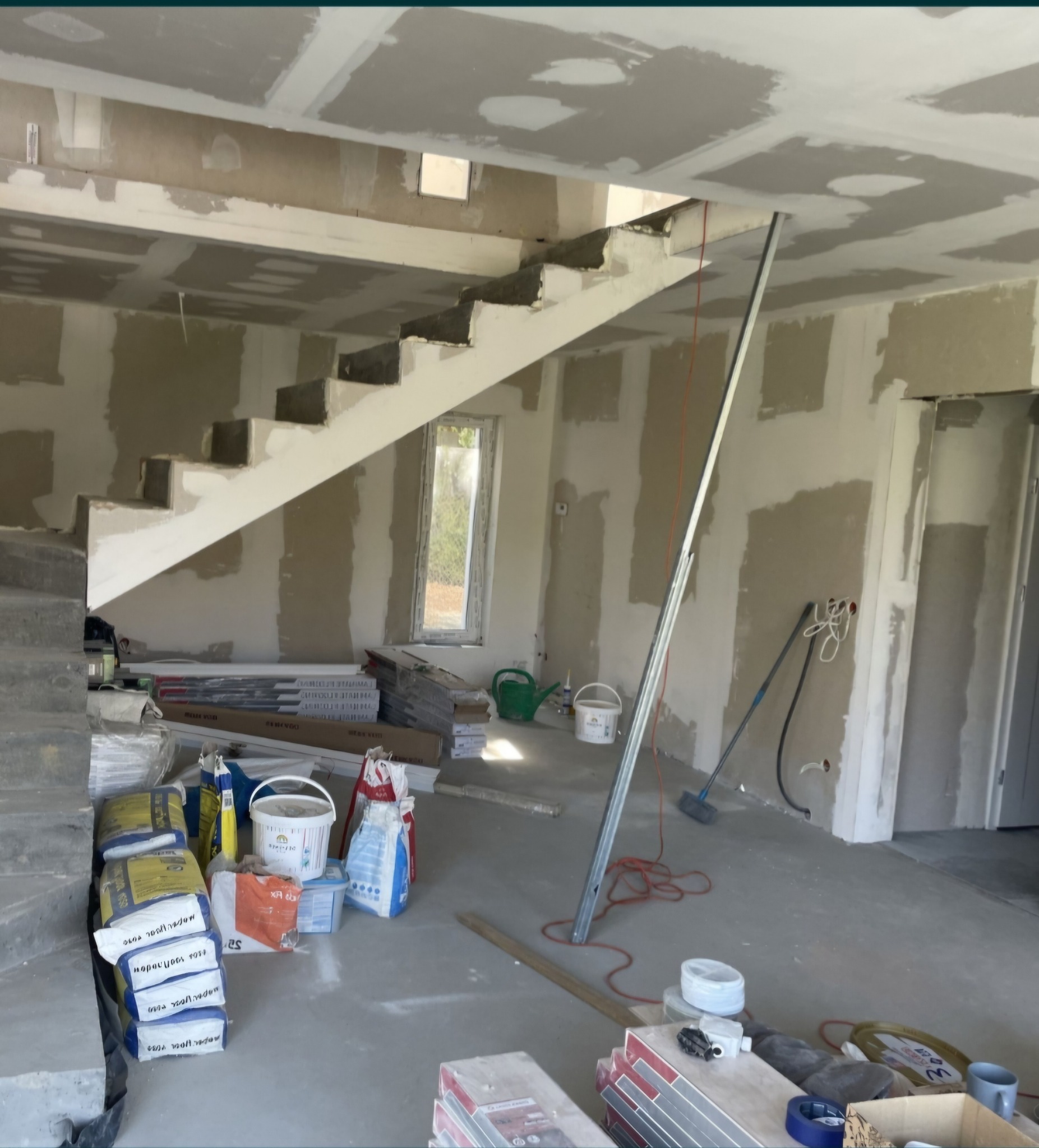 Wnętrze w trakcie prac wykończeniowych: surowe schody, ściany przygotowane pod malowanie, częściowo pokryte płytami gipsowo-kartonowymi, widoczne worki z zaprawą, puszki z farbą i narzędzia budowlane.