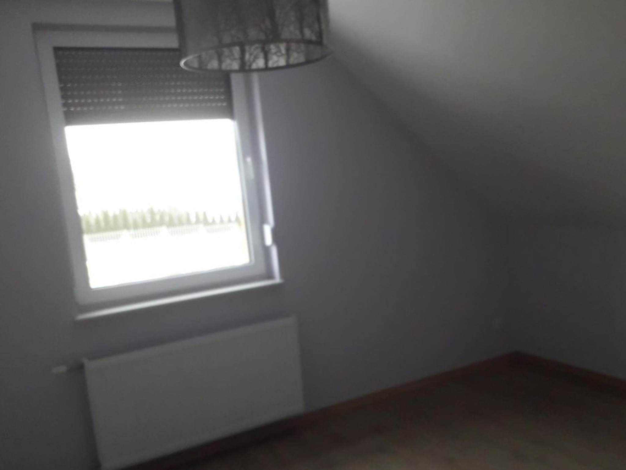 Poddasze ze skośną ścianą, świeżo pomalowane na szaro, z oknem z żaluzjami i grzejnikiem pod nim, widoczna lampa sufitowa.