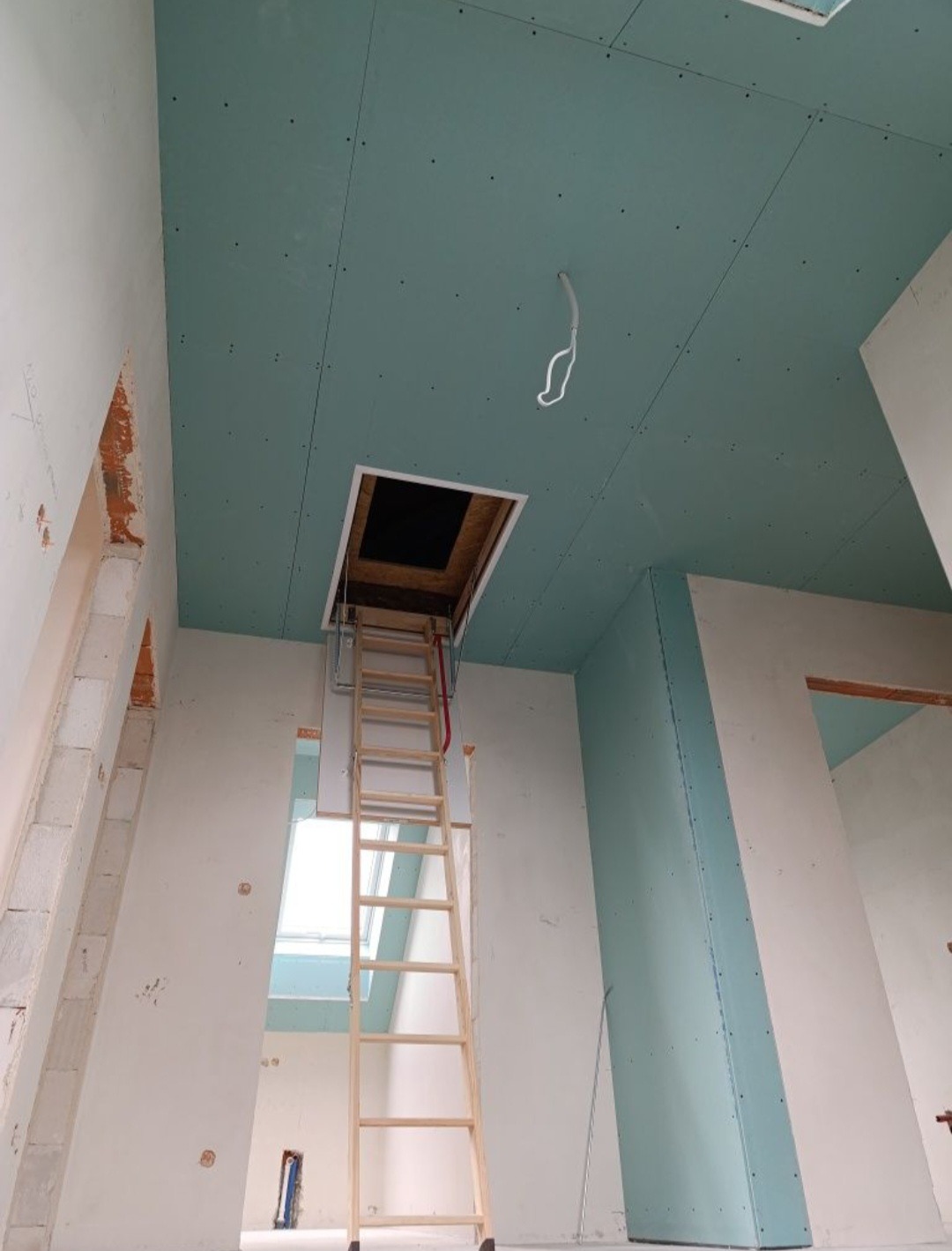 Montaż płyt kartonowo-gipsowych na suficie poddasza, widoczna drabina prowadząca do otworu strychowego, sufit w kolorze jasnej zieleni, ściany w trakcie wykończenia.