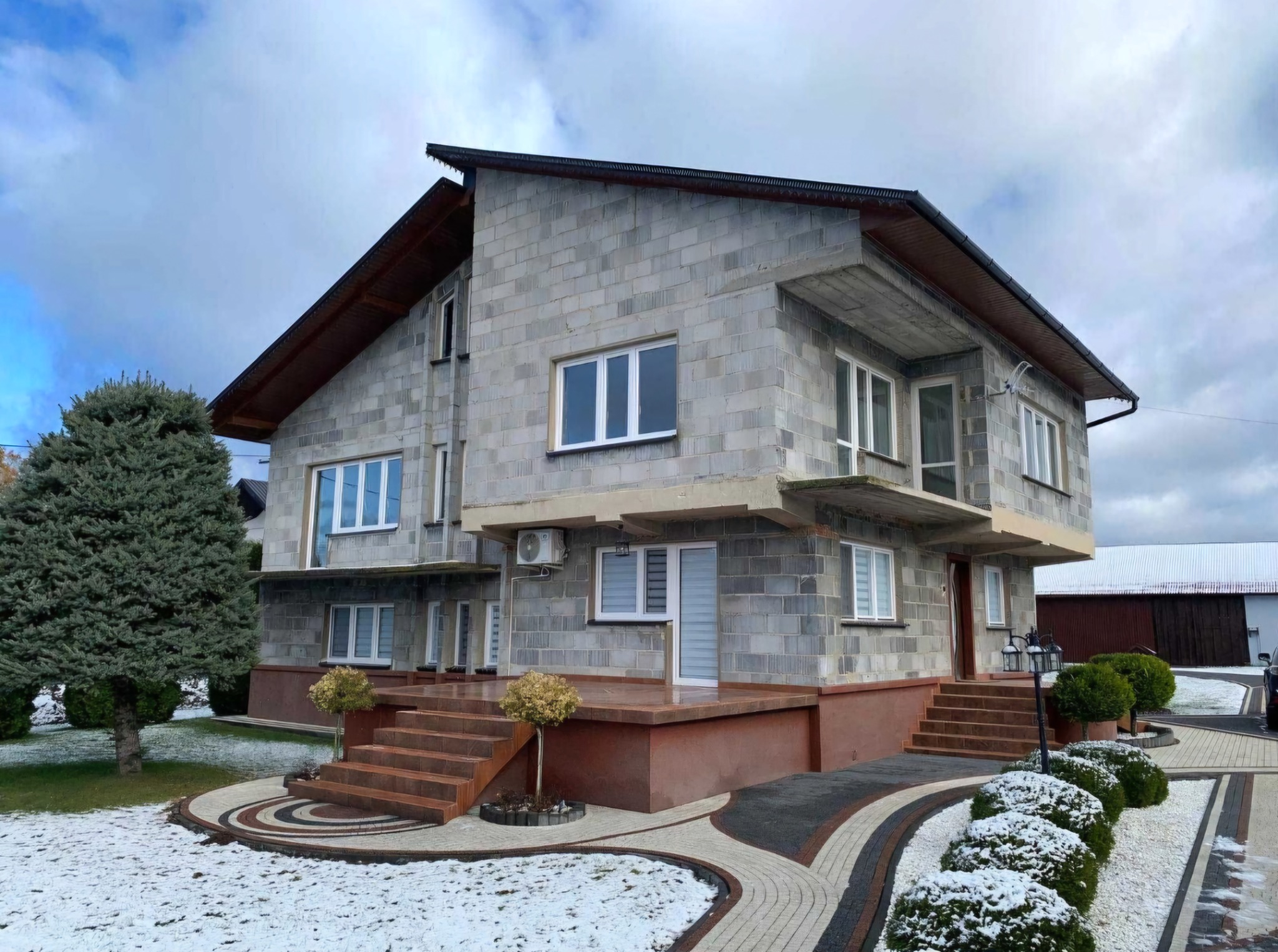 Surowy dwupiętrowy dom z szarego bloczka w trakcie budowy, z widocznymi elementami ocieplenia i częściowo wykończonym otoczeniem.