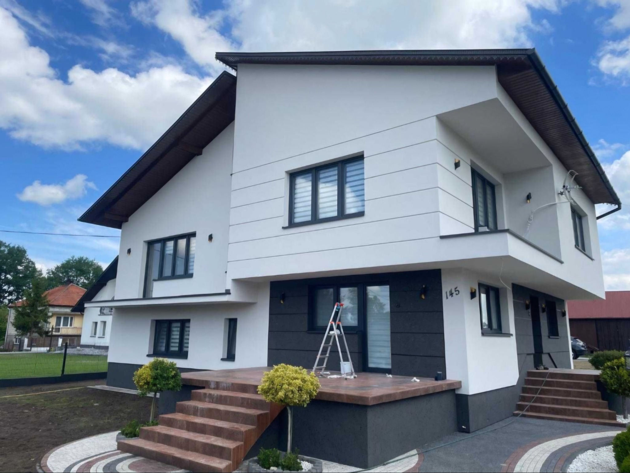 Nowoczesny dom jednorodzinny z białą elewacją, ciemnymi oknami i wejściem z brązowymi schodami, na których stoi aluminiowa drabina, numer domu 145.