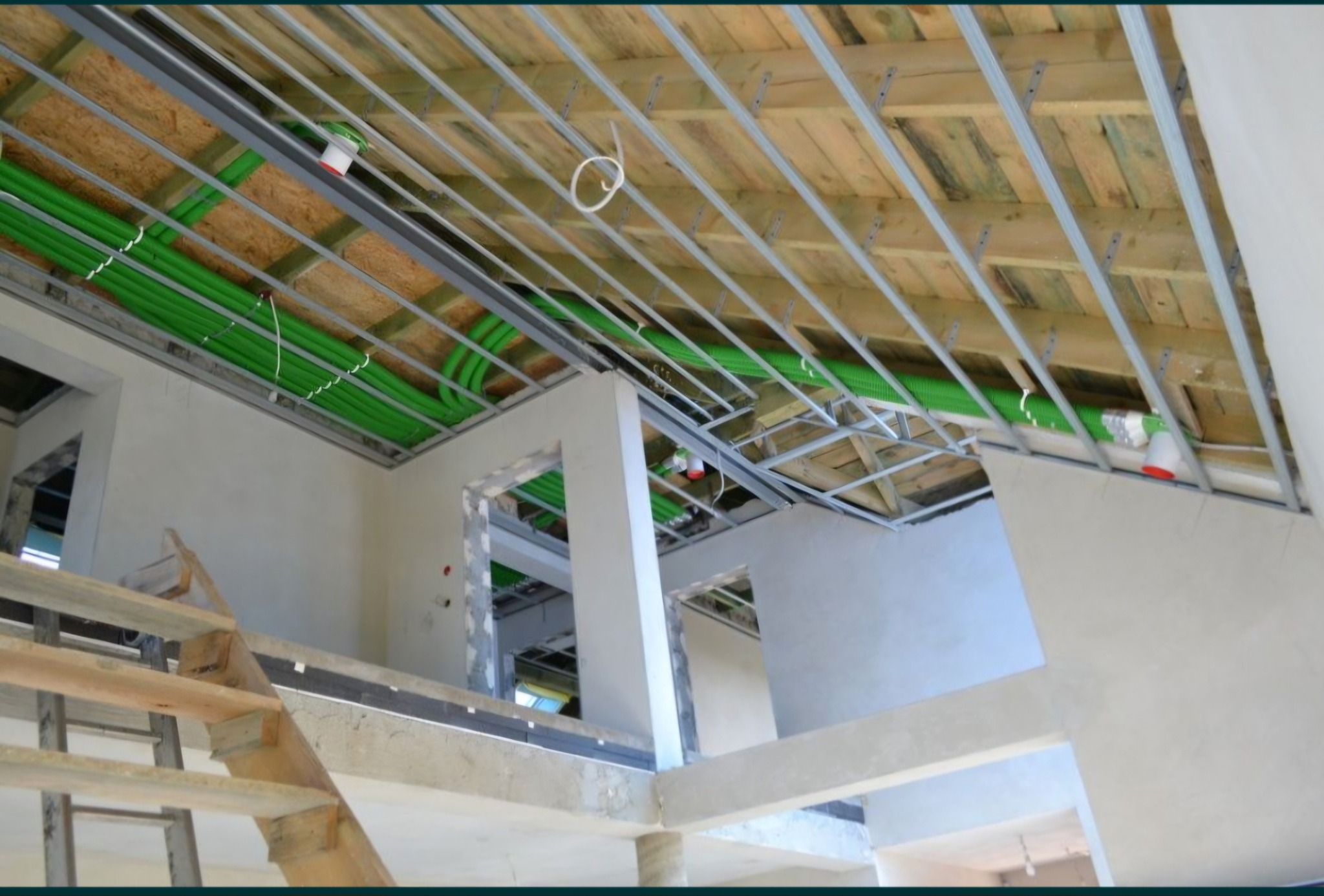 Wnętrze domu w trakcie remontu: widoczne drewniane poddasze z metalową konstrukcją, zielone rury instalacji, surowe ściany i fragment drewnianych schodów.