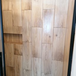 Usługi Budowlane Michał Hiller - Ściana w łazience wyłożona podłużnymi płytkami imitującymi jasne drewno, z wnęką pełniącą funkcję półki.