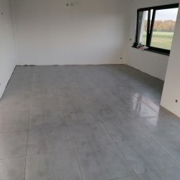 Usługi Budowlane Michał Hiller - Wykończone wnętrze z ułożoną szarą podłogą z płytek, widoczne białe ściany w trakcie przygotowania do malowania, okno z widokiem na zewnątrz i fragment schodów.