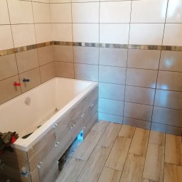 Usługi Budowlane Michał Hiller - Remontowana łazienka z częściowo wyłożonymi płytkami w odcieniach beżu i brązu, widoczna wanna obudowana płytkami, na wannie leżą robocze rękawice, a na ścianach i podłodze krzyżyki dystansowe.