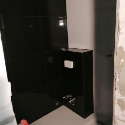 Usługi Budowlane Michał Hiller - Częściowo wykończona łazienka z czarnymi, błyszczącymi płytkami na ścianie i zabudowie stelaża WC, jasne płytki podłogowe, widoczne fragmenty surowego muru i instalacji hydraulicznej.