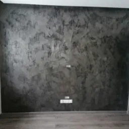 Ściana pokryta dekoracyjną, ciemnoszarą gładzią w stylu betonu architektonicznego, z widocznymi gniazdkami elektrycznymi i włącznikami, oraz fragmentem okna i drzwi.