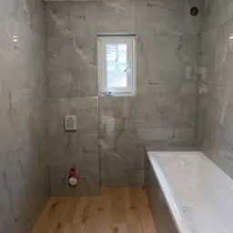 Wykładanie szarych, marmurkowych płytek w łazience z drewnopodobną podłogą, widoczne przyłącza hydrauliczne, okno i wanna zabezpieczona folią.