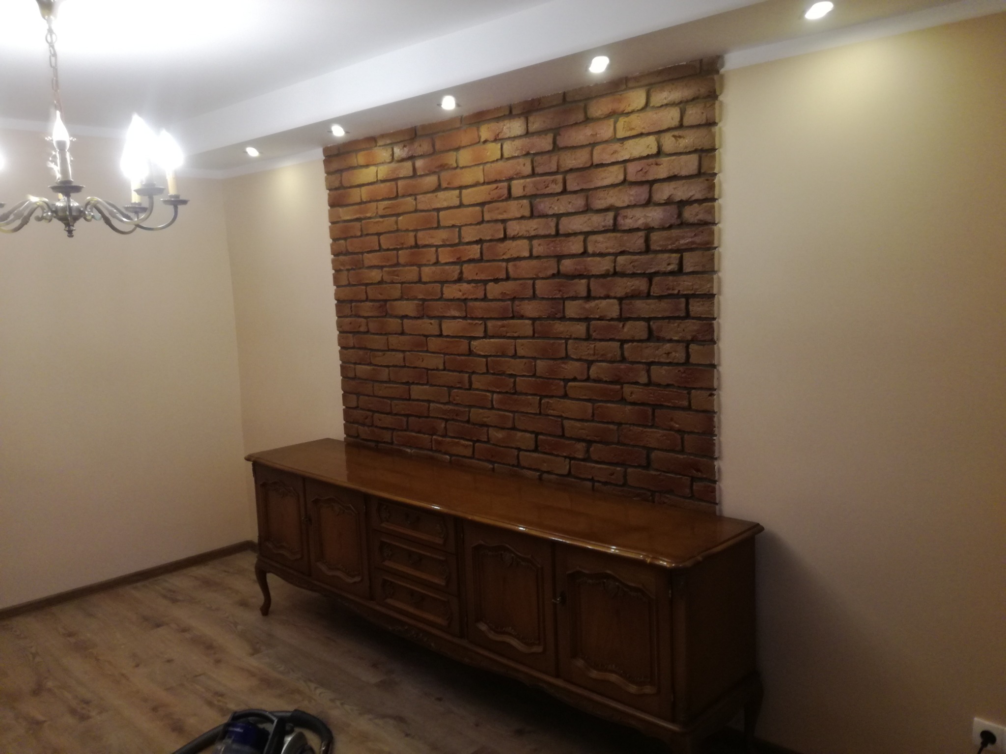 Salon z dekoracyjną ścianą z cegły, oświetleniem punktowym i antycznym kredensem z ciemnego drewna.