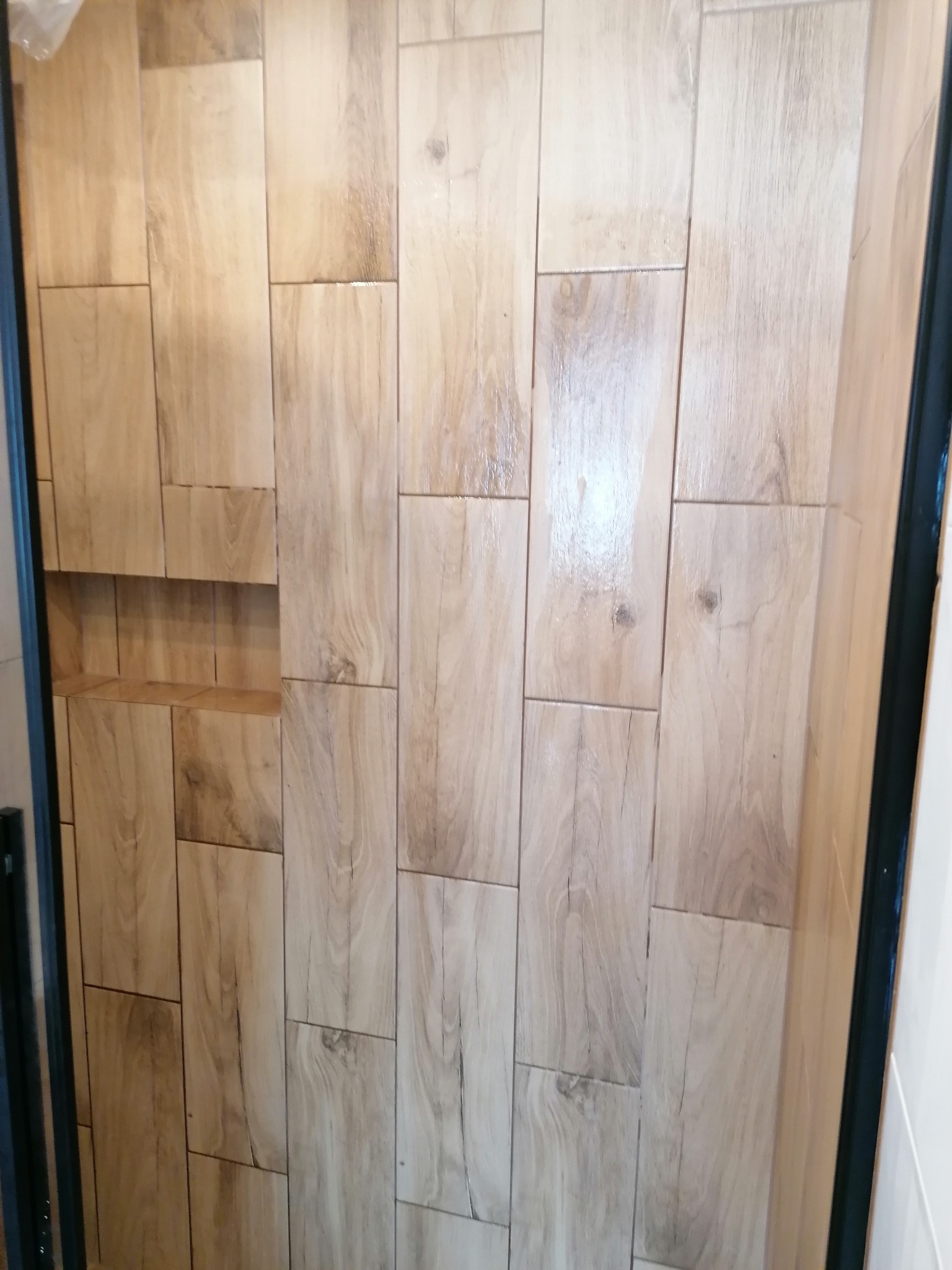 Ściana w łazience wyłożona podłużnymi płytkami imitującymi jasne drewno, z wnęką pełniącą funkcję półki.