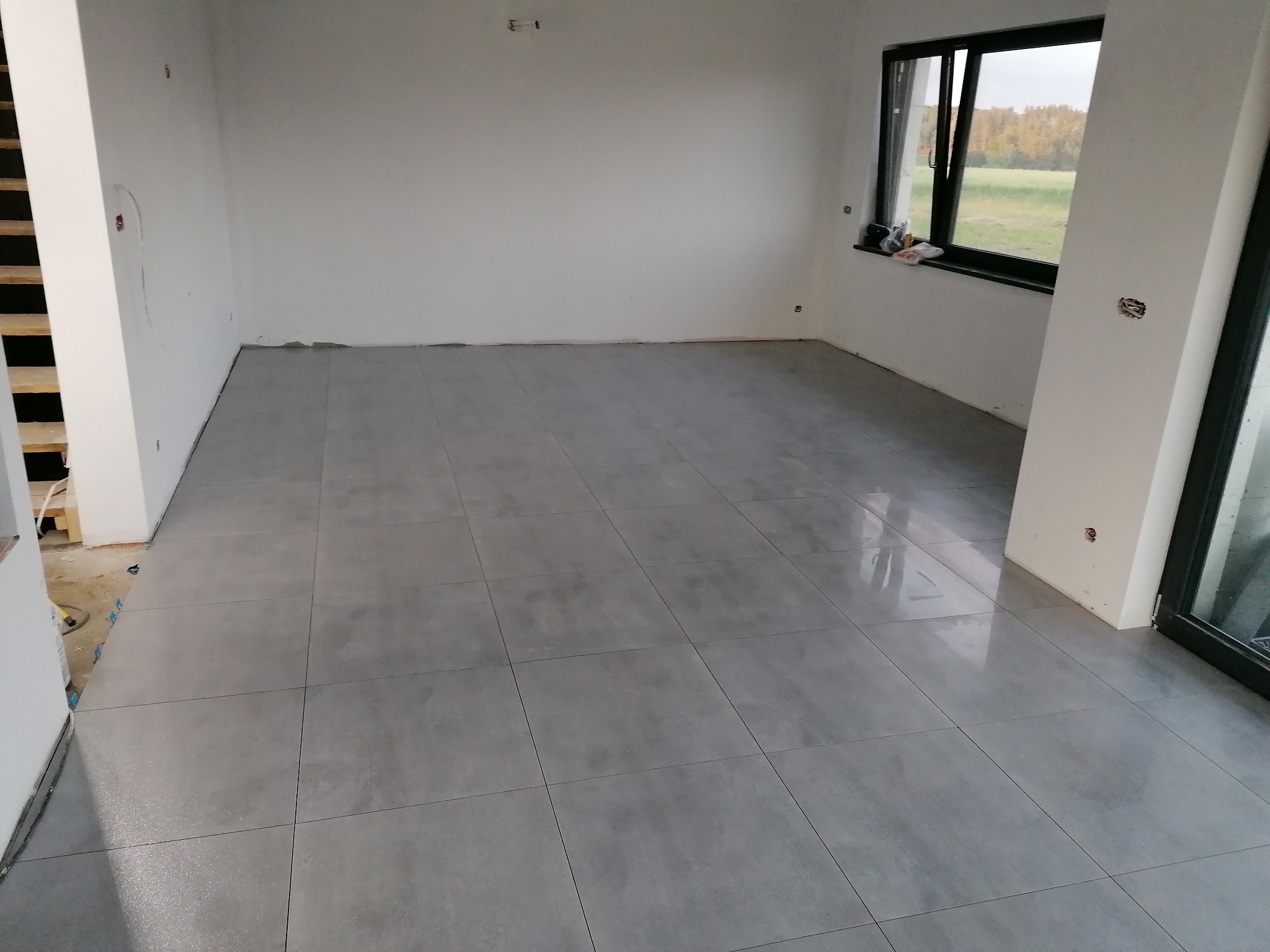 Wykończone wnętrze z ułożoną szarą podłogą z płytek, widoczne białe ściany w trakcie przygotowania do malowania, okno z widokiem na zewnątrz i fragment schodów.