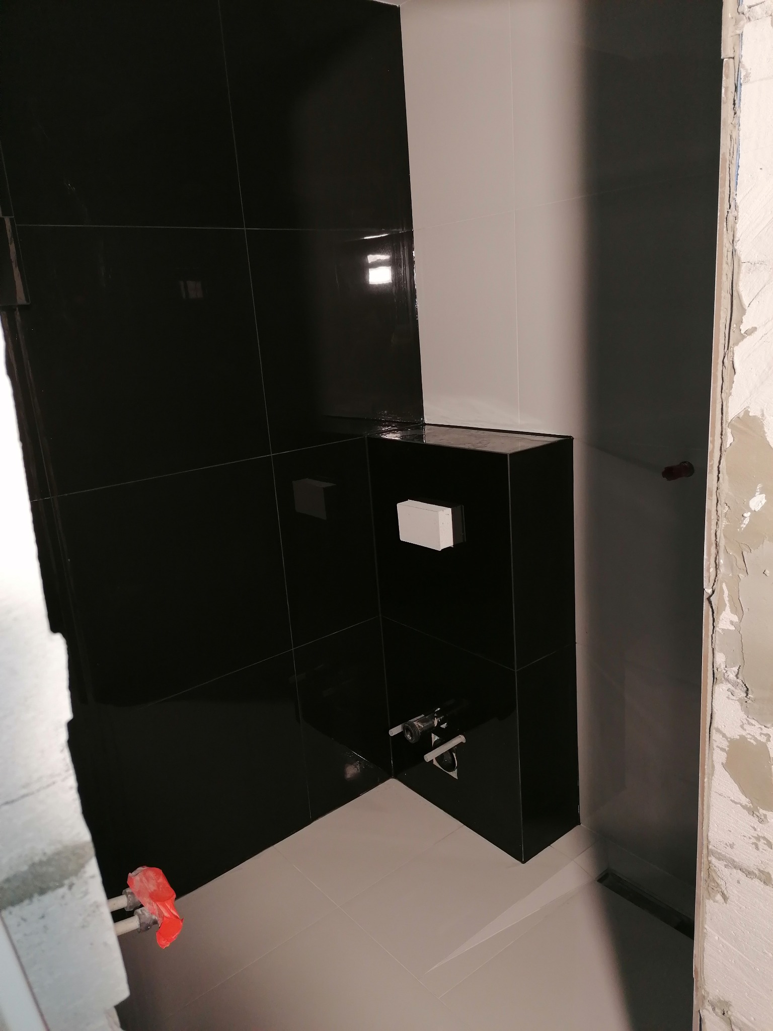 Częściowo wykończona łazienka z czarnymi, błyszczącymi płytkami na ścianie i zabudowie stelaża WC, jasne płytki podłogowe, widoczne fragmenty surowego muru i instalacji hydraulicznej.
