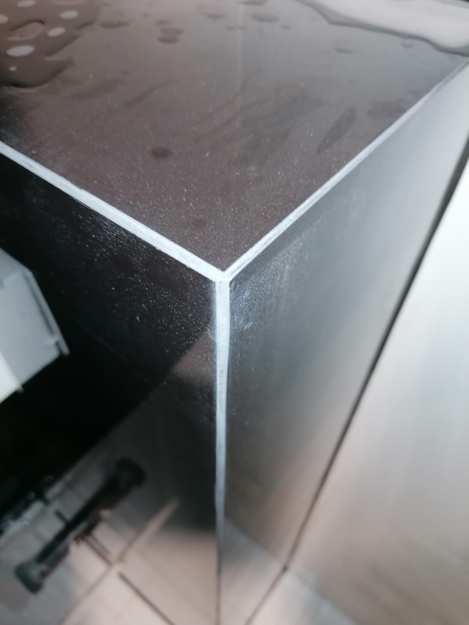 Czarny, błyszczący blat z widocznymi śladami wody i białym silikonem w narożniku, sfotografowany z bliska pod kątem.