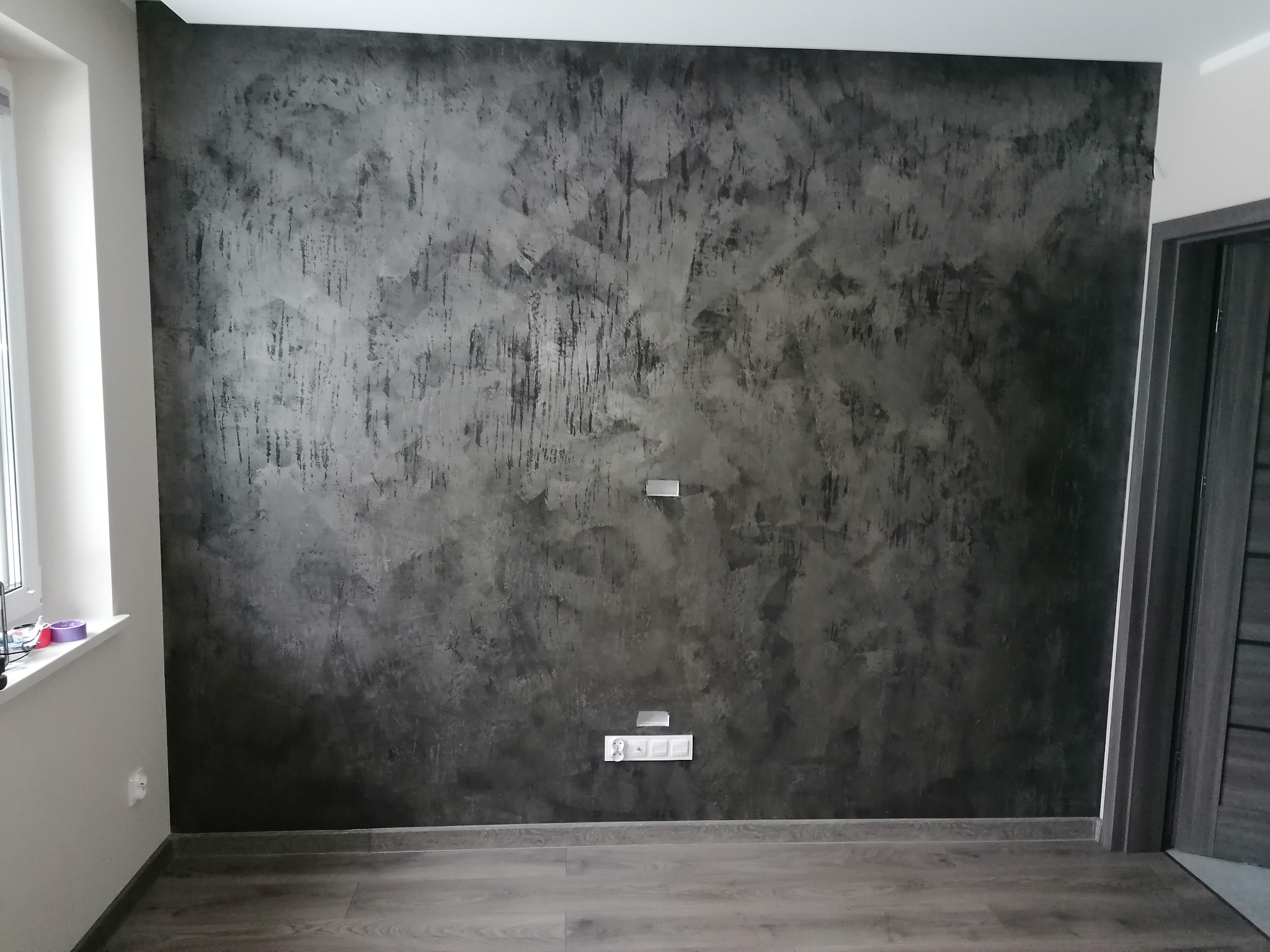 Ściana pokryta dekoracyjną, ciemnoszarą gładzią w stylu betonu architektonicznego, z widocznymi gniazdkami elektrycznymi i włącznikami, oraz fragmentem okna i drzwi.