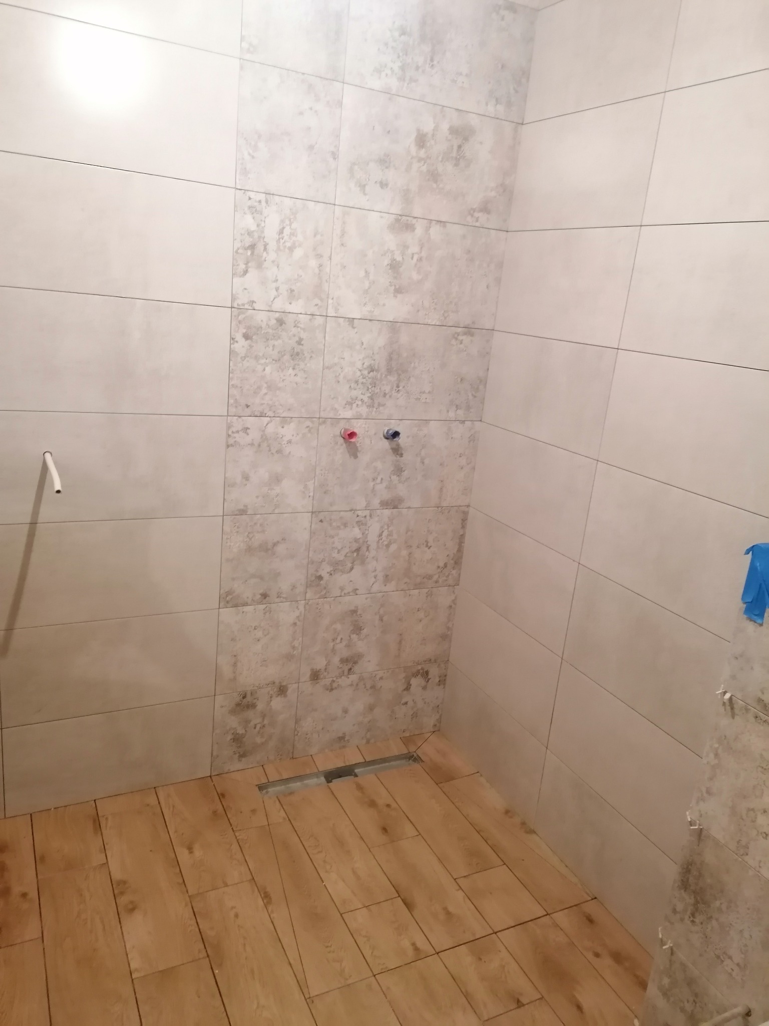 Wnętrze łazienki w trakcie remontu, widoczne płytki ścienne w odcieniach beżu i szarości, imitujące kamień, drewnopodobna podłoga, odpływ liniowy w podłodze, wystające rurki instalacji wodnej...