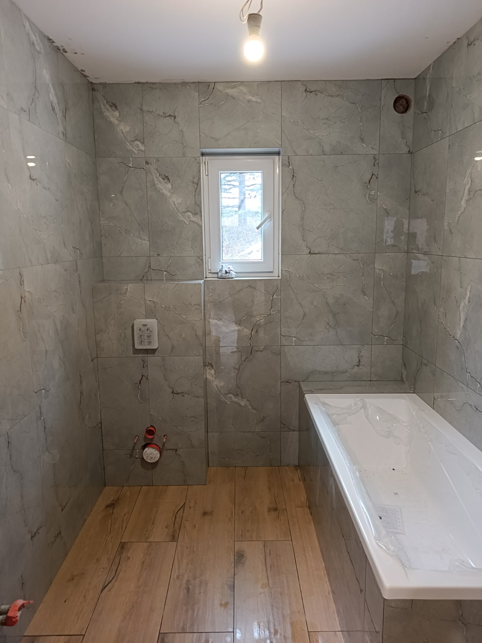 Wykładanie szarych, marmurkowych płytek w łazience z drewnopodobną podłogą, widoczne przyłącza hydrauliczne, okno i wanna zabezpieczona folią.